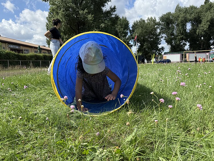 Sportfest der 4- bis 6-jährigen Kinder Ein Kind mit Kappe kriecht durch einen blauen Spieltunnel auf einer grünen Wiese, während im Hintergrund Erwachsene und andere Kinder stehen.