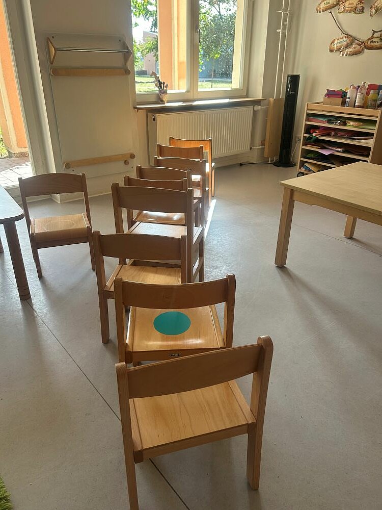 Heller Gruppenraum mit niedrigen Holzstühlen, die aufgereiht hintereinanderstehen und einem Tisch. Im Hintergrund ist ein Regal mit Materialien, ein Fenster mit Vorhang und ein Heizkörper. Einer der Stühle trägt einen grünen Punkt als Markierung auf der Sitzfläche – ein typischer Gruppenraum der Kita.