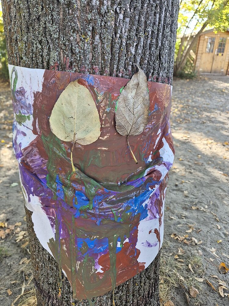 Ein Baumstamm mit einem bunt bemalten Blatt Papier, das mit grüner, blauer, roter und brauner Farbe gestaltet wurde. Zwei trockene Blätter sind oben auf das Papier gelegt und bilden die Augen eines Gesichtes. Im Hintergrund sind ein sandiger Boden, einige gelbe Blätter und ein Holzgebäude zu sehen.