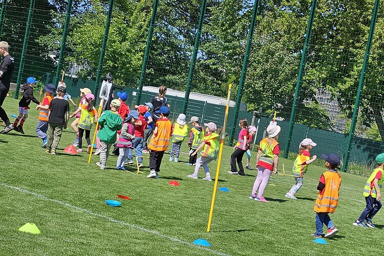Festspiele mit Ritter Keule beim 1. FC Union Berlin Eine große Gruppe von Kindern mit Sicherheitswesten und Sonnenhüten laufen auf einem Sportfeld scheinbar wild durcheinander. Auf dem Rasen liegen bunte Kegel und es stecken gelbe Markierungen im Boden. Es sind auch zwei Erwachsene zu sehen.