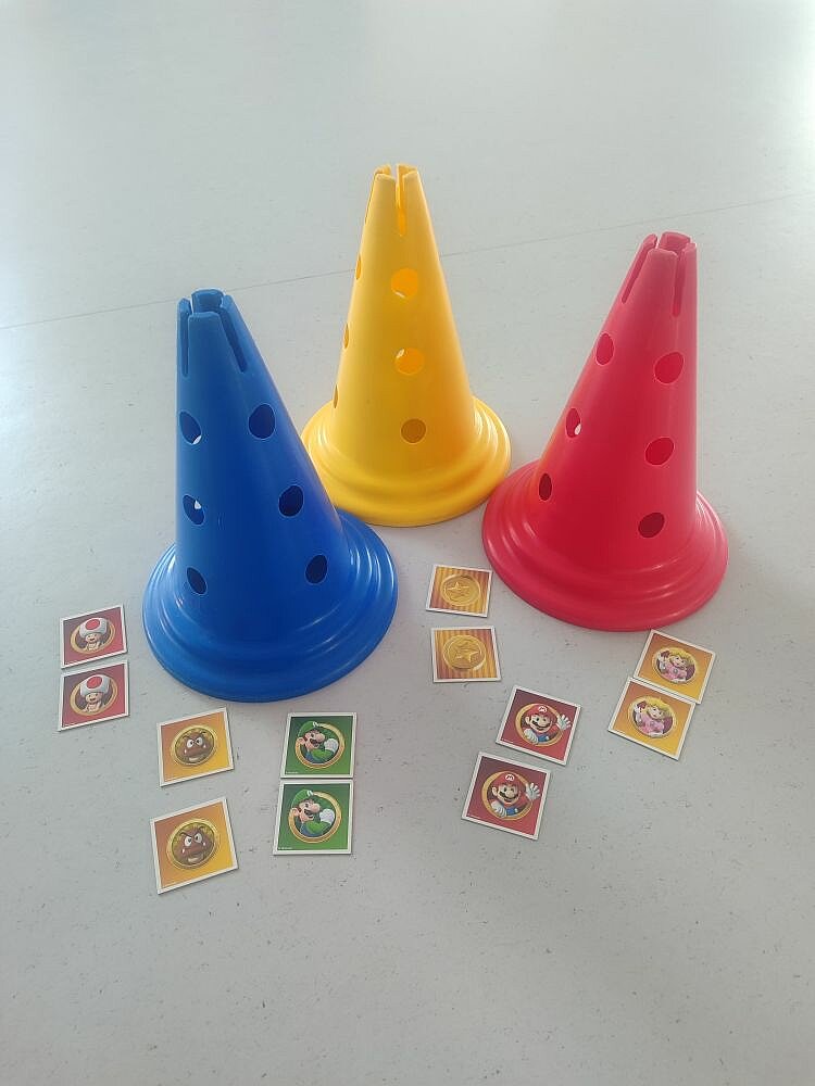 Drei bunte Plastik-Hütchen in Blau, Gelb und Rot mit Löchern, umgeben von kleinen runden Aufklebern mit Motiven aus dem Mario-Spiel, wie Pilzen, Sternen und Charakteren wie Mario und Luigi.
