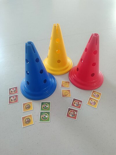 Drei bunte Plastik-Hütchen in Blau, Gelb und Rot mit Löchern, umgeben von kleinen runden Aufklebern mit Motiven aus dem Mario-Spiel, wie Pilzen, Sternen und Charakteren wie Mario und Luigi.