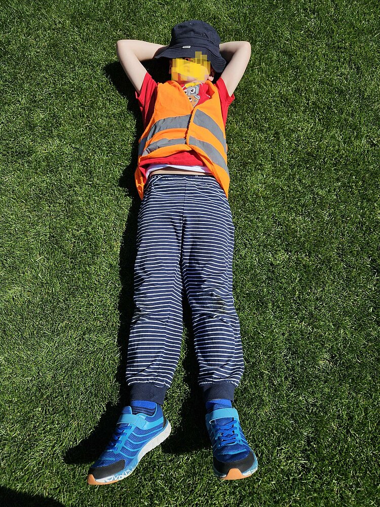 Festspiele mit Ritter Keule beim 1. FC Union Berlin Ein Kind liegt auf dem Rücken auf einem grünen Feld und trägt eine orangefarbenes Sicherheitsweste, eine blaue Hose, blaue Turnschuhe und einen blauen Sonnenhut..