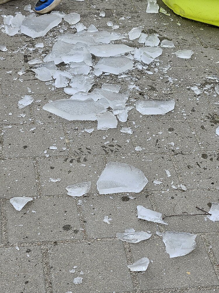 Auf einem grauen Steinboden liegen zerbrochene Eisschollen, am Rand ist ein blauer Kinderschuh zu sehen.