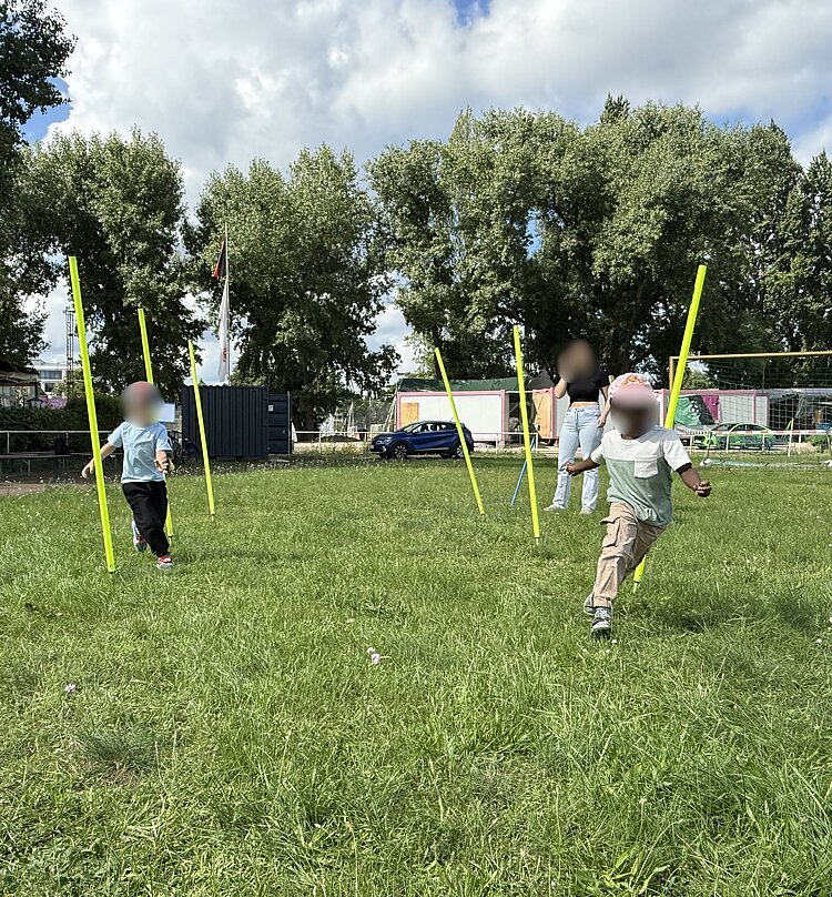 Sportfest der 4- bis 6-jährigen Kinder Kinder laufen zwischen aufgestellten gelben Stangen hindurch auf einer Wiese, während Erwachsene im Hintergrund zuschauen.