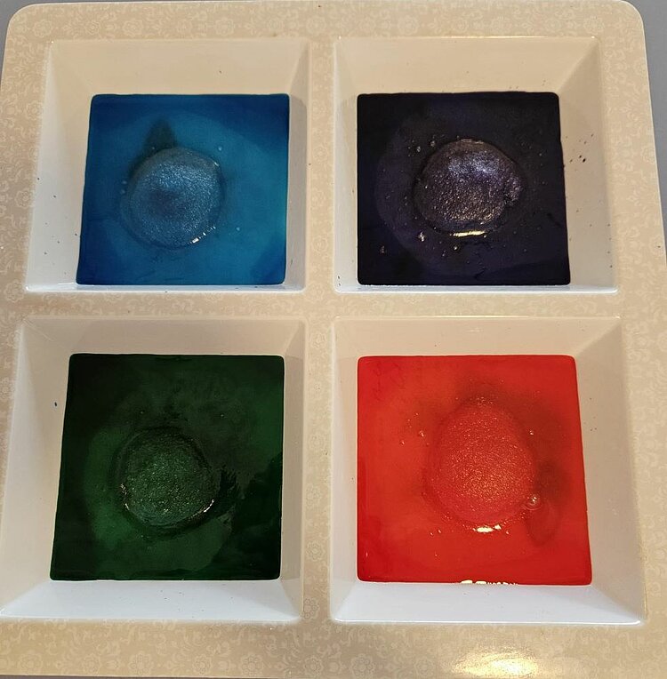 Eine helle Palette mit vier quadratischen Vertiefungen, gefüllt mit verschiedenen Farben: oben links blau mit einem blauen Farbklecks in der Mitte, oben rechts dunkelblau/schwarz mit einem dunklen Farbklecks, unten links grün mit einem grünen Farbklecks und unten rechts rot mit einem roten Farbklecks.