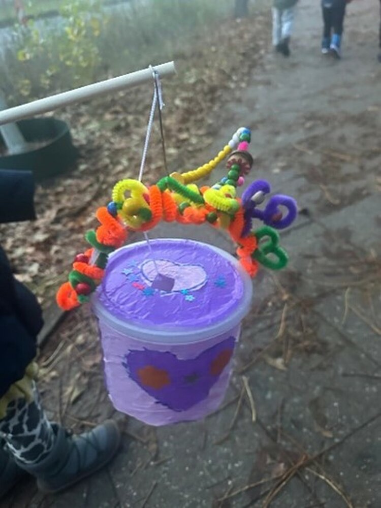 Eine selbstgebastelte Laterne aus einem lila Plastikbecher, der mit bunten, gewundenen Pfeifenreinigern und kleinen Pompons verziert ist. Der Becher ist an einem Stock befestigt und hängt im Freien. Im Hintergrund sind ein Weg und ein paar Personen zu sehen.