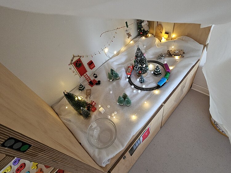Liebevoll gestaltete Winterlandschaft mit Mini-Tannenbäumen, Lichterkette, Spielzeugeisenbahn und Schneedekoration auf einem Podest im Gruppenraum.