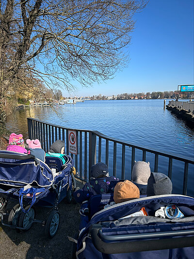 Zwei Kinderwägen mit mehreren Kindern stehen an einem blauen Gewässer vor einem Geländer; im Hintergrund Bäume und ein Steg.