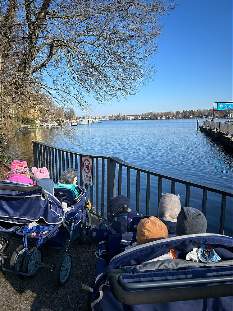 Zwei Kinderwägen mit mehreren Kindern stehen an einem blauen Gewässer vor einem Geländer; im Hintergrund Bäume und ein Steg.