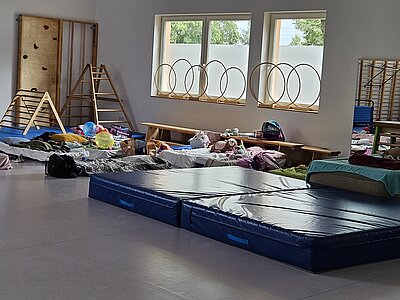 Ein unvergessliches Kita-Abenteuer: Unsere Übernachtung voller Magie und Gemeinschaft Im Sportraum der KITA Adlerküken liegen mehrere Matratzen mit Decken und Schlafsäcken darauf. Außerdem stehen dort Koffer, Taschen Kuscheltiere der Übernachtungskinder. Auf dem Fensterbrett stehen verschieden große Ringe aus Holz. Auch einen Wandspiegel kann man ganz rechts im Bild erkennen.