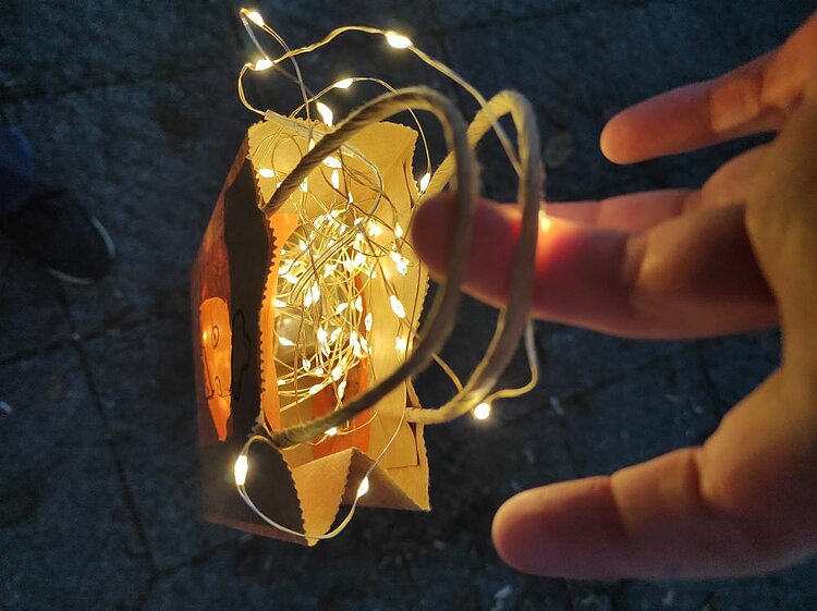 Eine Hand hält eine kleine Laterne aus durchsichtigem Papier, die mit goldenen Lichterketten gefüllt ist. Die Laterne ist mit einem Draht verschlossen und leuchtet warm in der Dunkelheit.