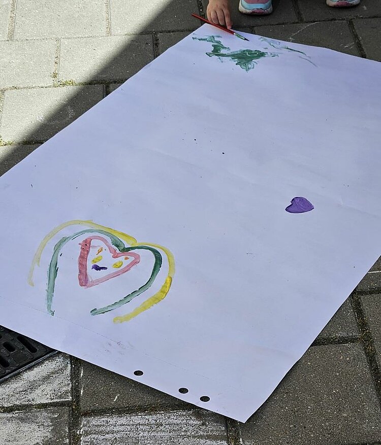 Ein Kind malt auf einem großen, weißen Blatt Papier auf dem Boden. Das Bild zeigt einen bunten Herzumriss, der mit gelben, grünen und roten Linien verziert ist. Im Hintergrund sind weitere Farbspuren in Grün und ein kleiner violetter Abdruck zu sehen. Das Kind hält einen Pinsel in der Hand.