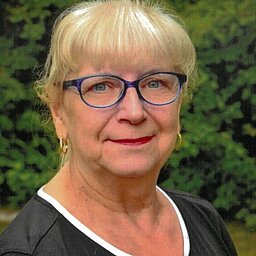 Zu sehen ist Frau Stange mit einem schwarzen Oberteil und einer blauen Brille, die in Nahaufnahme in die Kamera lächelt. Im Hintergrund sind Nadelbäume.