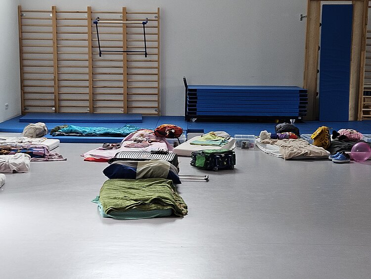 Ein unvergessliches Kita-Abenteuer: Unsere Übernachtung voller Magie und Gemeinschaft Im Sportraum der KITA Adlerküken liegen mehrere Matratzen mit Decken und Schlafsäcken darauf. Außerdem stehen dort Koffer und Taschen der Kinder. Am Rand sind blauen Matten vor den Sprossenwänden aufgebaut, auch ein ganzer Mattenwagen steht dort.