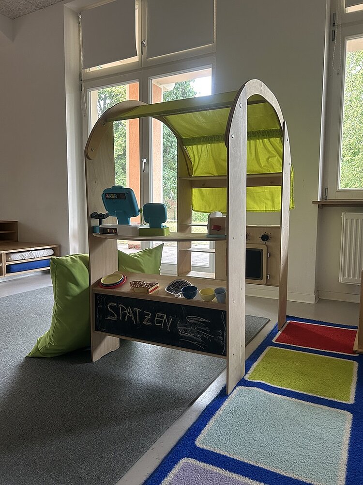 Im Vordergrund steht ein hölzernes Spielhaus mit gebogenem Dach und einer leuchtend gelben Stoffabdeckung. Auf den Regalböden sind verschiedene Spielzeuge und Gegenstände aus Holz und Kunststoff angeordnet. Vor dem Spielhaus befindet sich ein schwarzes Kreidebrett mit der Aufschrift 'SPATZEN' und einer einfachen Zeichnung. Rechts liegt ein bunter Teppich mit quadratischen Feldern in Rot, Gelb, Blau und Grün. Im Hintergrund sind große Fenster mit Blick ins Grüne sowie ein Heizkörper und weitere Möbelstücke zu sehen.