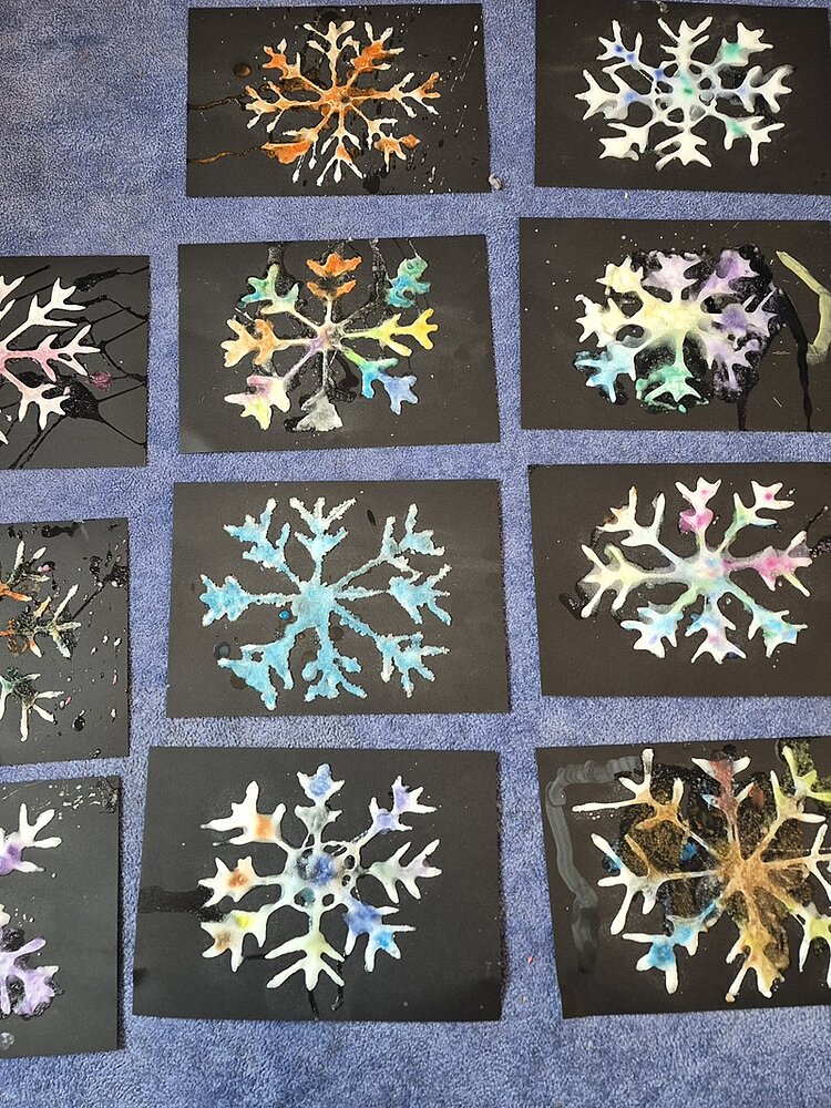 Farbenfrohes Schneeflockendesign auf schwarzem Papier. Die Schneeflocken sind in leuchtenden Farben wie Orange, Blau, Lila und Grün bemalt und bilden einen auffälligen Kontrast zum dunklen Hintergrund. Das Arrangement sieht kreativ und optisch ansprechend aus und lässt auf eine spielerische und künstlerische Aktivität schließen.