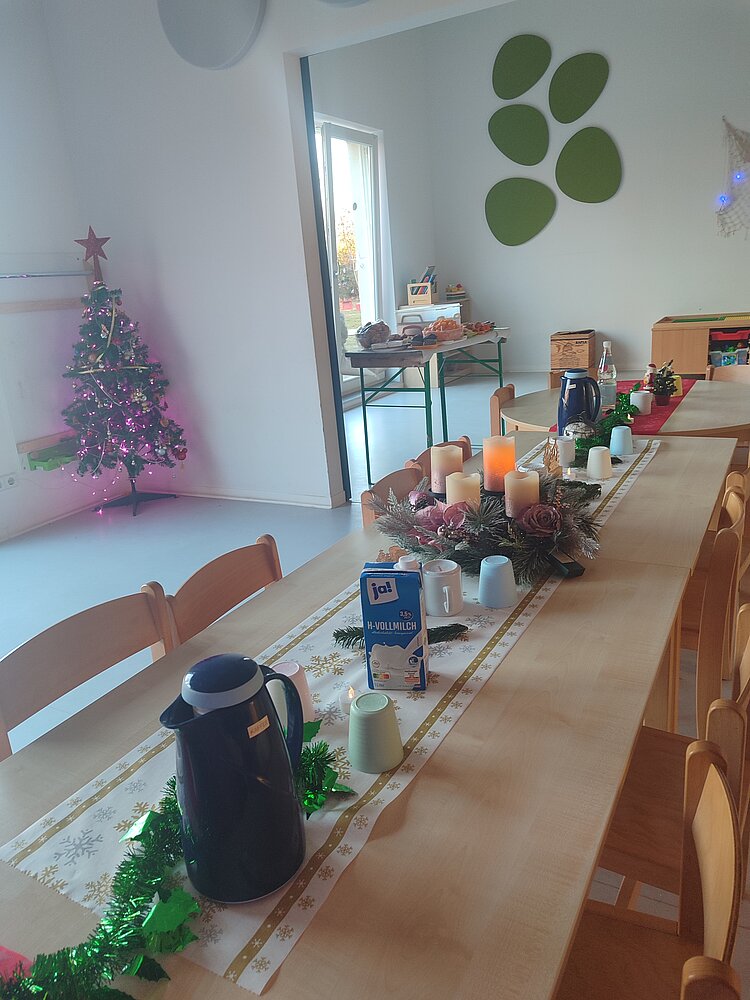 Festlich geschmückter Kita-Raum mit langer Kaffeetafel, Adventsgesteck mit Kerzen und im Hintergrund ein leuchtender Weihnachtsbaum neben einem Buffettisch.