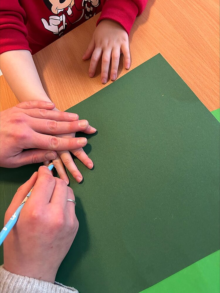 Eine Nahaufnahme zeigt die Hände eines Kindes und einer Erzieherin auf einem Holztisch. Das Kind trägt einen roten Pullover mit einem Mickey-Mouse-Motiv. Die erwachsene Person hält einen hellblauen Buntstift und zeichnet den Umriss der Hand des Kindes auf ein dunkelgrünes Blatt Papier. Die Szene spielt in der KITA Adlerküken, wo Kinder und Erzieher gemeinsam weihnachtliche Dekorationen anfertigen. Das gezeichnete Handumrissmotiv soll später ausgeschnitten werden und Teil eines selbst gebastelten Tannenbaumes werden, bei dem die ausgeschnittenen Handumrisse der Kinder die Tannenzweige darstellen sollen.