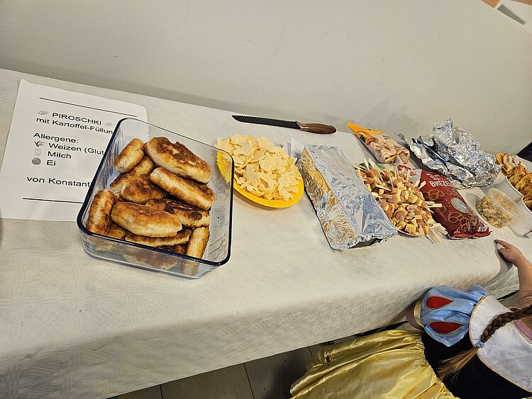 Buffet-Tisch mit einer Glasform voller gebratener Piroschki (Kartoffelfüllung), daneben Chips, eingepackte Speisen und kleine Snackspieße; links liegt ein Schild mit Allergenen.