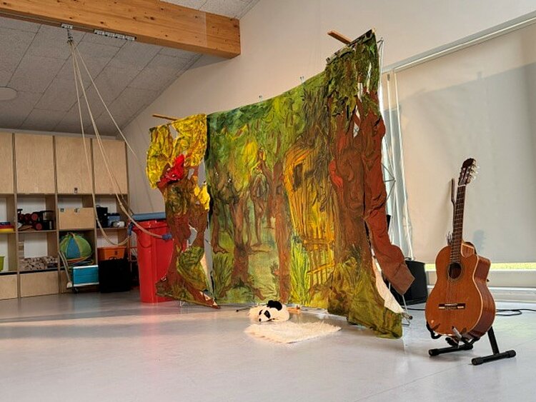 In einem großen, hellen Raum steht eine mit Waldmotiven bemalte Bühnenwand, davor liegen ein weißes Fell und ein schwarz-weißer Bärenkopf; rechts daneben steht eine Gitarre.