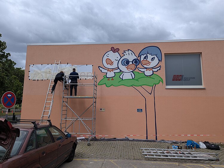 Wir können uns sehen lassen! Eine Hauswand mit einem Graffiti. Davor zwei Männer auf einem Gerüst, die ein Logo neben dem Graffiti anbringen.