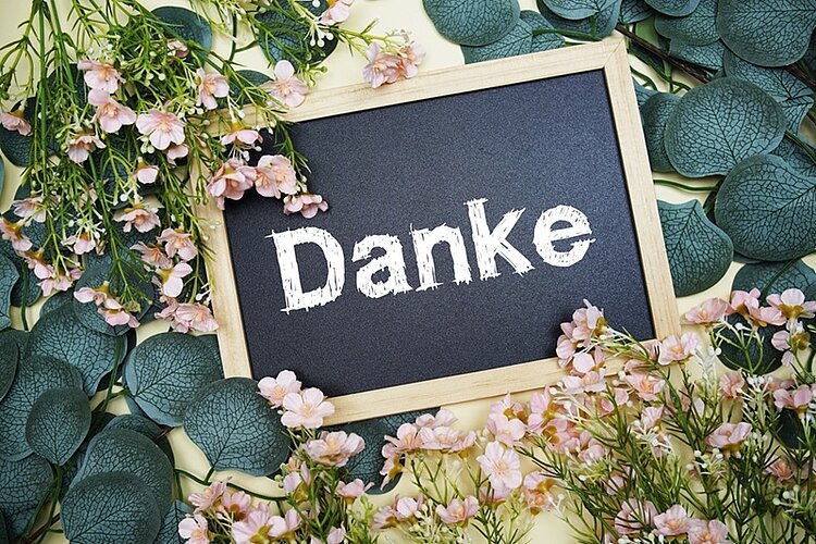 Eine kleine Tafel mit dem weißen Schriftzug 'Danke' liegt auf einem Untergrund, der mit grünen Blättern und zarten rosa Blüten bedeckt ist. Die Tafel hat einen hellen Holzrahmen.