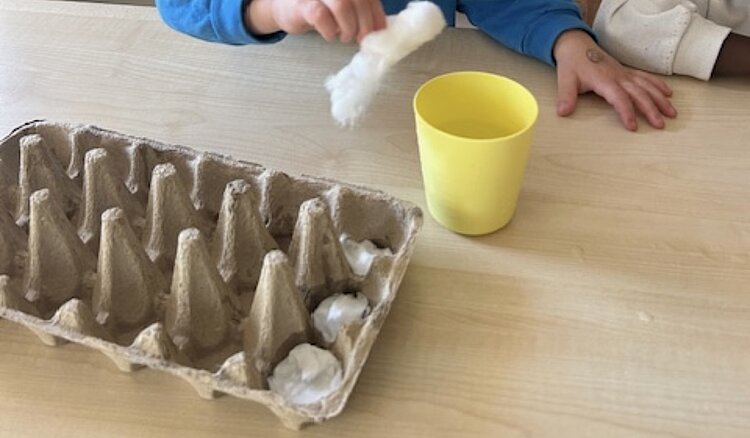Kinder gestalten an einem Tisch kleine Kugeln aus Watte oder Stoff; daneben stehen ein gelber Becher und ein Eierkarton.
