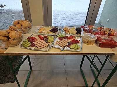Buffet-Tisch vor großen Fenstern mit Brötchen in Schalen, Tellern mit Wurst, Käse, Gurkenscheiben und Paprika sowie Trauben; daneben Salzbrezeln, ein Salat/Dip und Gebäck, draußen liegt Schnee.