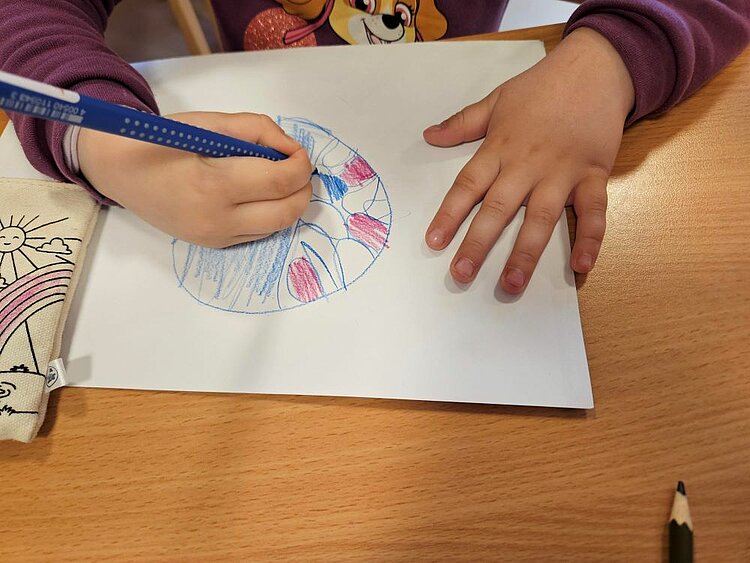 Es sind Kinderhände auf einem Tisch zu sehen. Eine Hand hält einen blauen Stift, mit dem ein Bild auf einem weißen Blatt Papier gemalt wird. Ein weiterer Stift und ein Mäppchen mit Sonnenmotiv liegen daneben.