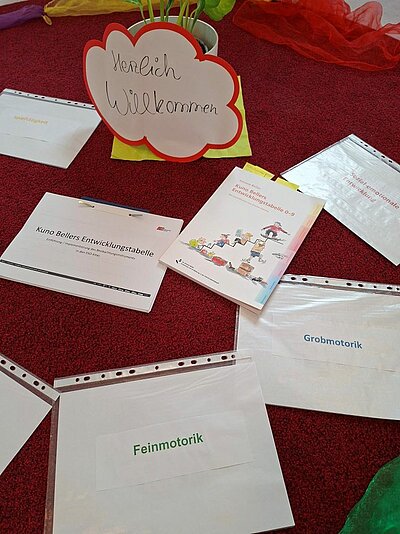 Ein Tisch mit roter Tischdecke auf dem verschiedenen Unterlagen für ein Seminar liegen. Zu sehen sind mehrere Papiere in Folien oder Hefter mit Beschriftungen wie „Kuno Bellers Entwicklungstabelle“, „Sozial-emotionale Entwicklung“, „Grobmotorik“ und „Feinmotorik“. Daneben liegt an einen Blumentopf gelehnt eine aus Papier ausgeschnittene Wolke mit rotem Rand mit der Aufschrift „Herzlich Willkommen“.