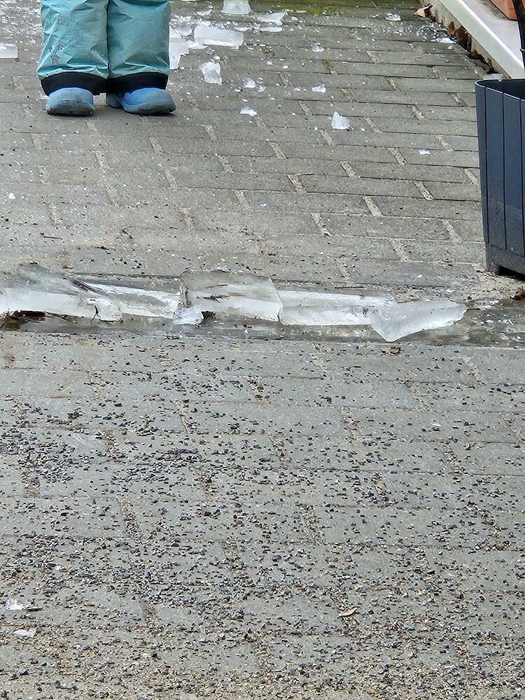 Auf einem grauen Steinboden liegen Eisstücke und Kies; am oberen Bildrand sieht man Kinderfüße in Stiefeln und türkisfarbener Hose.