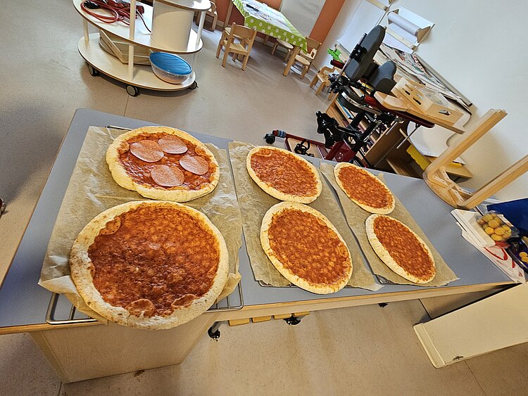 Ein unvergessliches Kita-Abenteuer: Unsere Übernachtung voller Magie und Gemeinschaft Auf einem Tisch liegen sechs große, runde Pizzen, die mit Tomatensoße belegt sind, eine davon schon mit Salami. Im Hintergrund befindet sich ein Schreibtisch ein kleiner Tisch mit winzigen Stühlen für die Kinder.