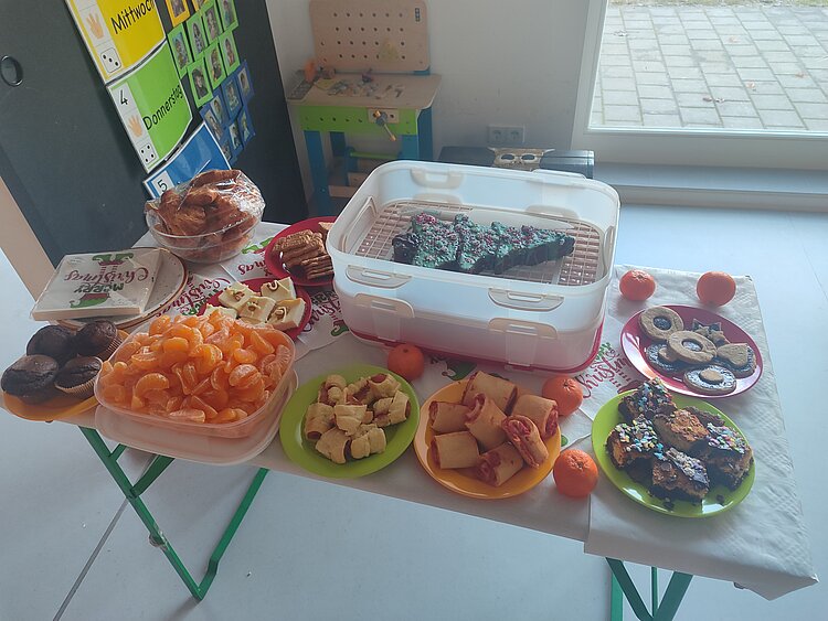 Bunt bestückter Buffettisch in der Kita mit Mandarinen, Gebäck, Muffins, Kuchenstücken und herzhaften Snacks auf farbigen Tellern.
