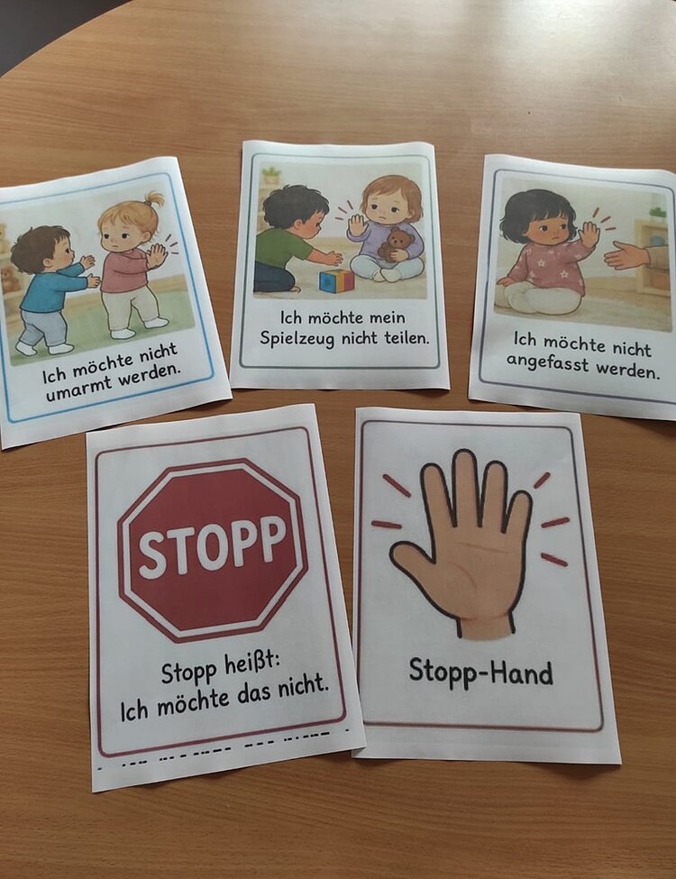 Fünf Karten liegen auf einer Holzoberfläche. Drei Karten zeigen kindliche Illustrationen: Ein Kind mit blonden Haaren und ein Kind mit braunen Haaren stehen sich gegenüber, daneben steht 'Ich möchte nicht umarmt werden'. Auf der zweiten Karte hält ein Kind sein Spielzeug fest, während ein anderes Kind daneben steht, mit dem Text 'Ich möchte mein Spielzeug nicht teilen'. Die dritte Karte zeigt ein Kind mit dunklen Haaren, das die Hände vor sich hält, mit dem Text 'Ich möchte nicht angefasst werden'. Die vierte Karte zeigt ein rotes Stoppschild mit dem Text 'STOPP heißt: Ich möchte das nicht.' Die fünfte Karte zeigt eine erhobene Hand mit dem Text 'Stopp-Hand'.