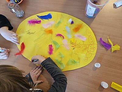 Auf einem Holztisch gestalten mehrere Kinder gemeinsam ein großes, gelbes, ovales Papier als Osterdekoration. Das Papier ist mit bunten Federn, Filzstiften und Klebstoff dekoriert. Kinderhände sind beim Malen und Basteln zu sehen. Auf dem Papier befinden sich bunte Kritzeleien und Zeichnungen.