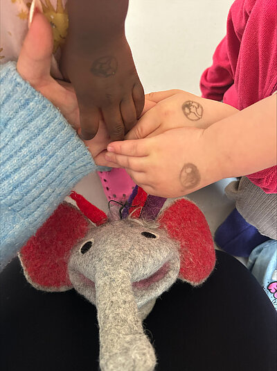 Kinder legen ihre Hände mit kleinen Stempelmotiven übereinander; darunter ist eine gefilzte Elefantenfigur zu sehen.