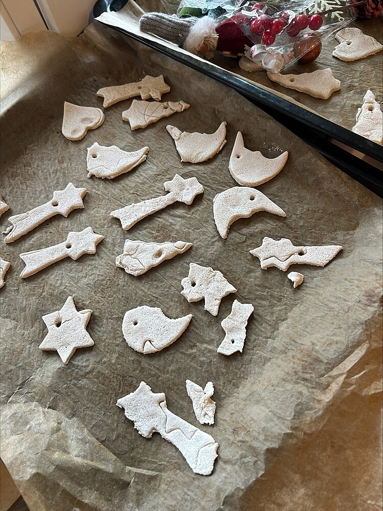 Auf einem mit Backpapier ausgelegten Backblech liegen verschiedene ungebackene Plätzchen in weihnachtlichen Formen, darunter Sterne, Herzen, Tannenbäume, Vögel, Engel und Glocken. Die Plätzchen sind hell und noch nicht gebacken. Im Hintergrund sind weihnachtliche Dekorationen wie rote Beeren und Stofffiguren sichtbar, die auf eine festliche Atmosphäre in der KITA Adlerküken hinweisen, wo Kinder und Erzieher gemeinsam Weihnachtsvorbereitungen treffen.