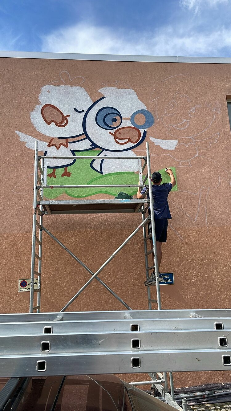 Wir können uns sehen lassen! Ein Mann, der auf einem Gerüst steht und auf eine Hauswand ein Graffiti in Form von Adlerküken in einem Nest sprüht.