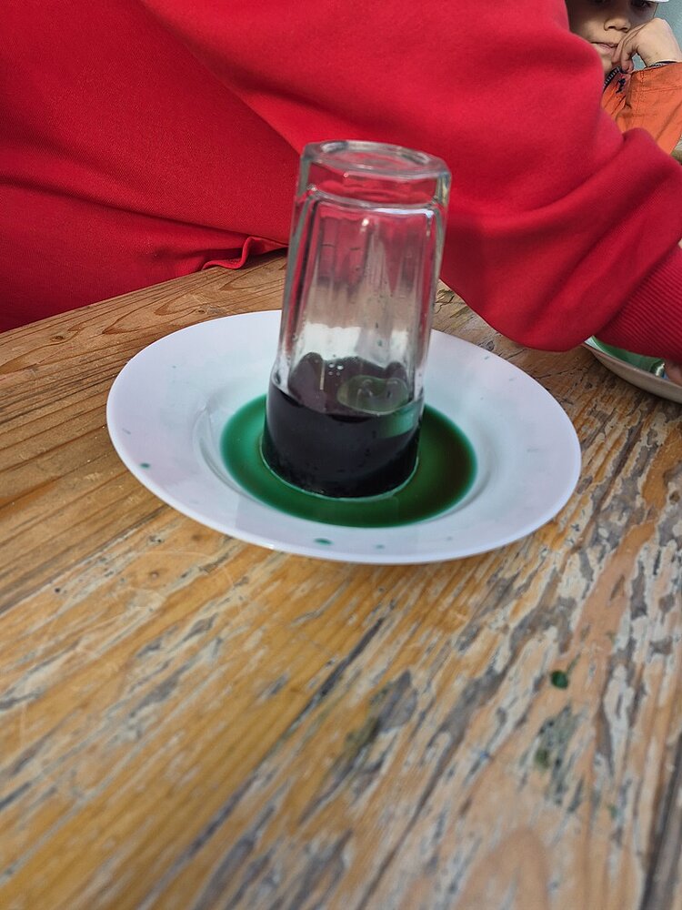Ein umgedrehtes Glas steht auf einem weißen Teller, aus dem sich eine grüne Flüssigkeit ringförmig ausbreitet. Der Holztisch zeigt deutliche Gebrauchsspuren. Im Hintergrund ist ein rotes Kleidungsstück zu sehen.