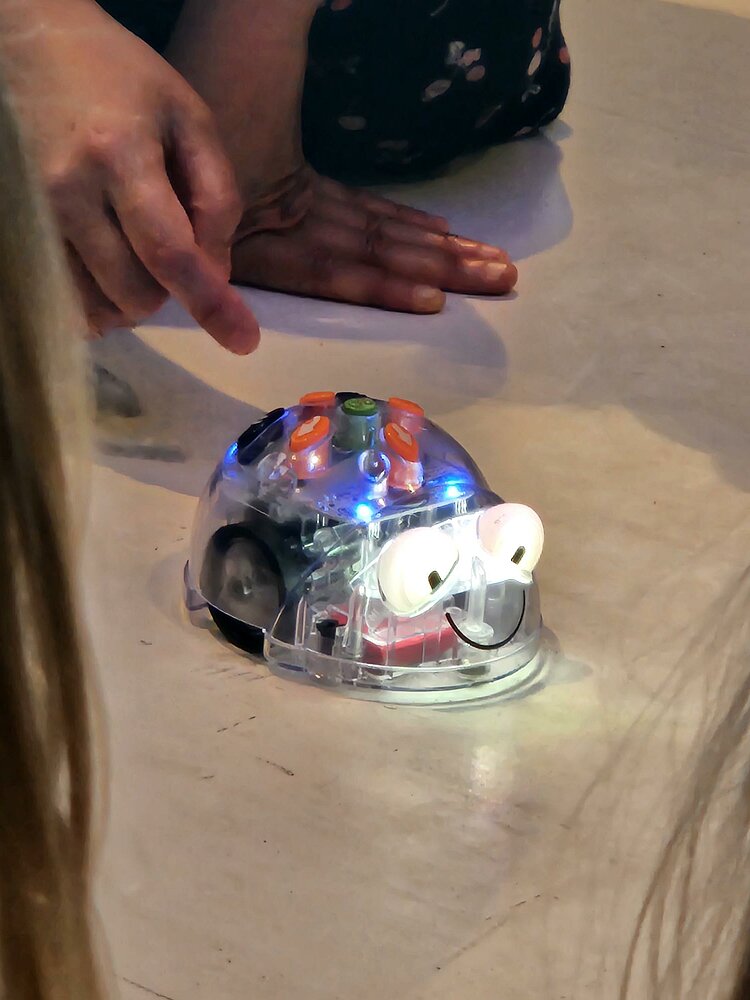 Eine Hand zeigt auf einen kleinen, käferförmigen Roboter mit bunten Lichtern, der auf dem Boden steht.