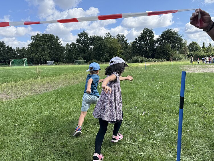 Sportfest der 4- bis 6-jährigen Kinder Zwei Kinder rennen lachend unter einem rot-weißen Absperrband hindurch, im Hintergrund ist eine Gruppe weiterer Kinder auf einer Wiese zu sehen.