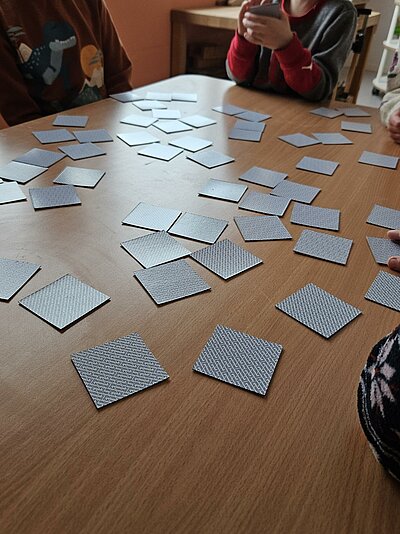 Auf einem Holztisch liegen viele verdeckte Memory-Karten; mehrere Personen sitzen darum und spielen gemeinsam.