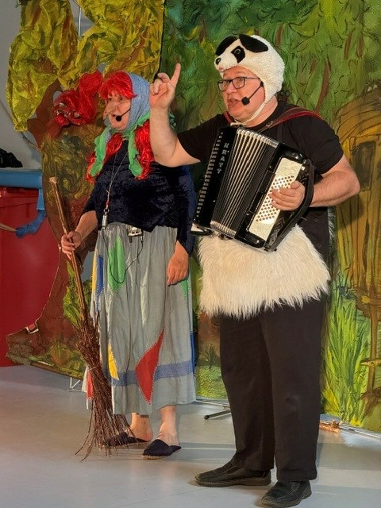 Vor einer bunt bemalten Wand stehen eine Frau im Hexenkostüm und ein Mann in einem schwarz-weißen Bärenkostüm mit Ziehharmonika. 