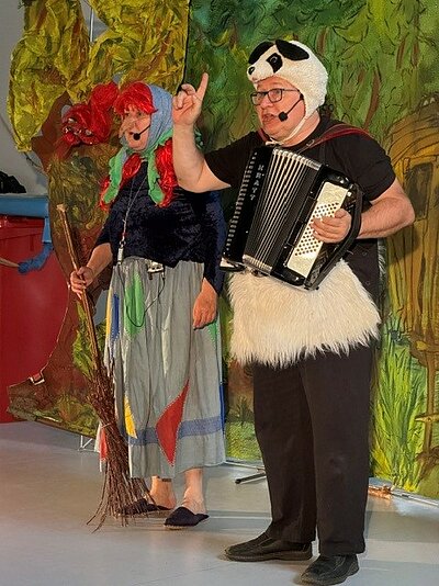Vor einer bunt bemalten Wand stehen eine Frau im Hexenkostüm und ein Mann in einem schwarz-weißen Bärenkostüm mit Ziehharmonika. 