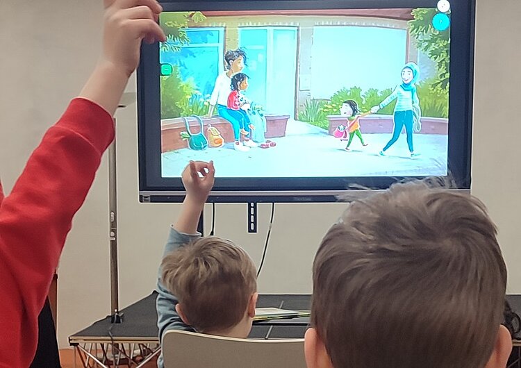 Drei Kinder sitzen in einem Raum und blicken auf einen großen Bildschirm, auf dem eine bunte Zeichentrickszene mit einer Familie im Garten gezeigt wird. Zwei Kinder heben die Hand, eines davon trägt einen roten Pullover. Die Szene auf dem Bildschirm zeigt zwei Erwachsene und zwei Kinder, die sich im Freien aufhalten. Im Hintergrund sind Pflanzen und ein Haus mit großen Fenstern zu sehen.