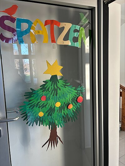 An einer Glastür in der KITA Adlerküken ist eine bunte Weihnachtsdekoration aus Papier angebracht. Oben steht in großen, farbigen Buchstaben das Wort 'SPATZEN'. Darunter ist ein Weihnachtsbaum aus grünen Papierhandabdrücken der Kinder gestaltet, die die Tannenzweige darstellen. Der Baum ist mit roten und gelben Papierkugeln geschmückt und trägt oben einen gelben Stern. Die Dekoration ist handgemacht und vermittelt eine festliche, kindgerechte und vorweihnachtlich besinnliche Atmosphäre.