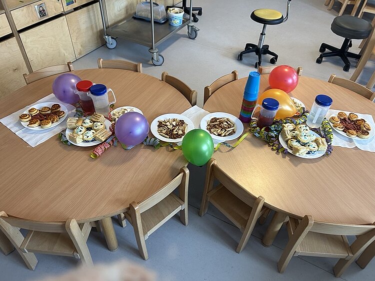 Zwei runde Holztische stehen nebeneinander, umgeben von kleinen Holzstühlen. Auf den Tischen liegen bunte Luftballons, Pappbecher, Servietten und mehrere Teller mit Gebäck, darunter Muffins und kleine Kuchenstücke. Die Tische sind festlich dekoriert, und im Hintergrund sind Regale und weitere Stühle zu sehen.