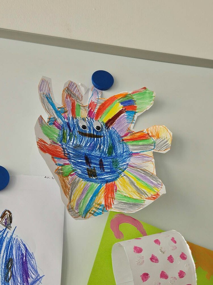 Eine Kinderzeichnung von einem bunten Fantasiewesen mit aufgeklebten Wackelaugen hängt an einer Magnettafel. Darunter sind weitere Kinderzeichnungen zu sehen.