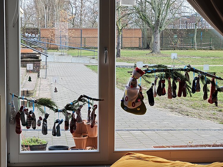 Socken-Adventskalender aus kleinen Socken, aufgehängt an Zweigen vor einem großen Fenster mit Blick in den Garten.